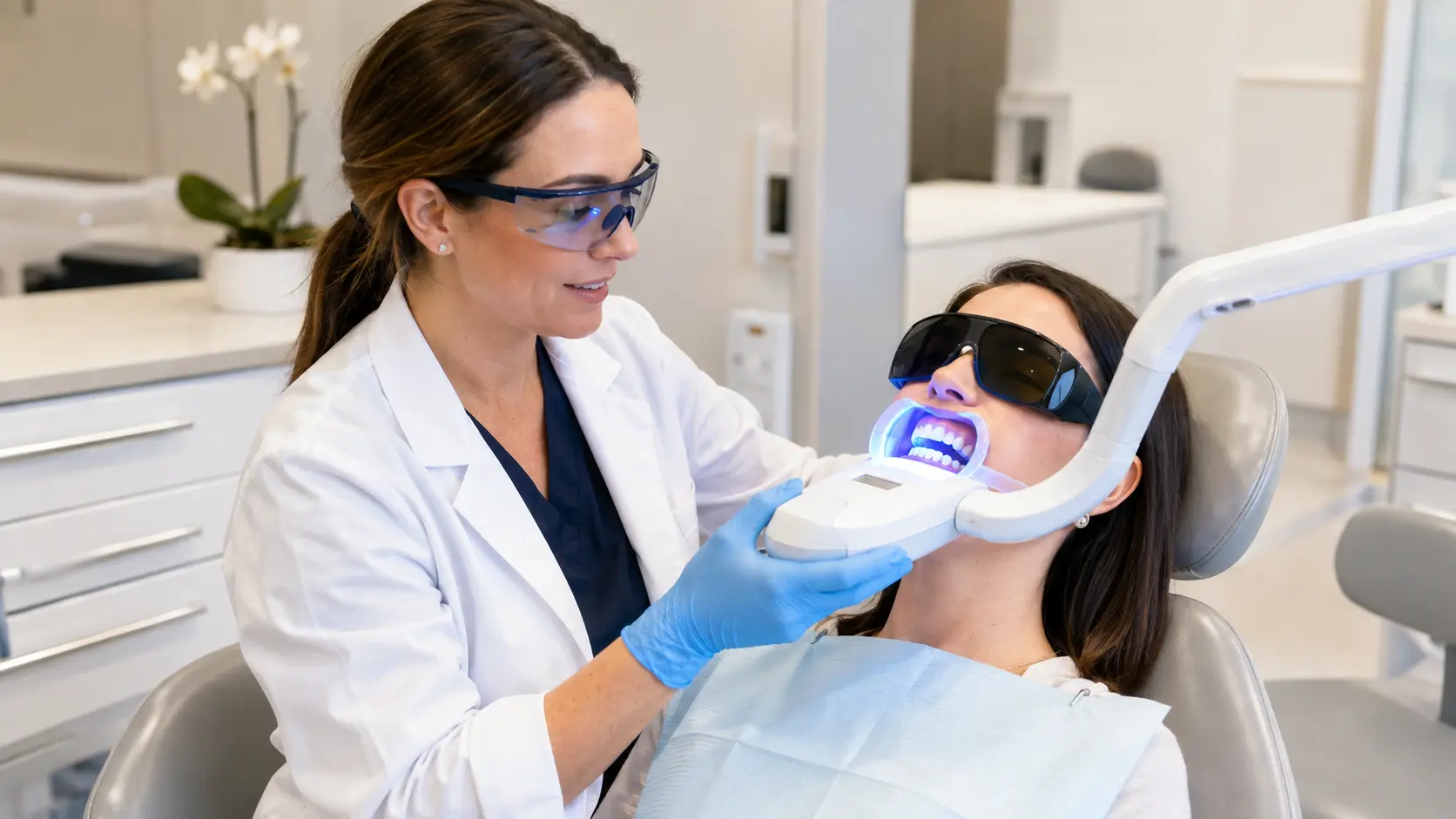 A dentist in Reston wearing protective eyewear and blue gloves performs an in-office teeth whitening treatment on a female patient, using a blue LED curing light to activate the bleaching gel.